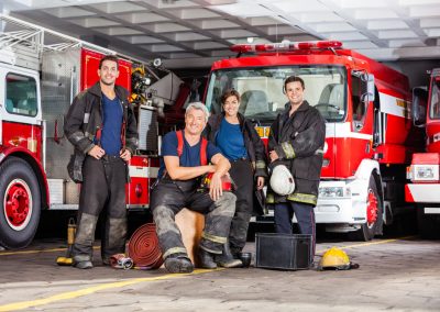 Portrait,Of,Happy,Firefighter’s,Team,With,Equipment,Against,Trucks,At
